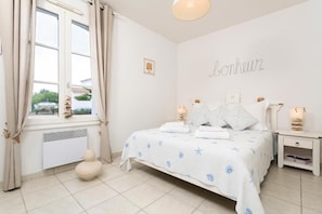 Une chambre à coucher comprenant un lit, des tables de chevet, une fenêtre avec des rideaux, un radiateur et un mur orné d’une affiche.