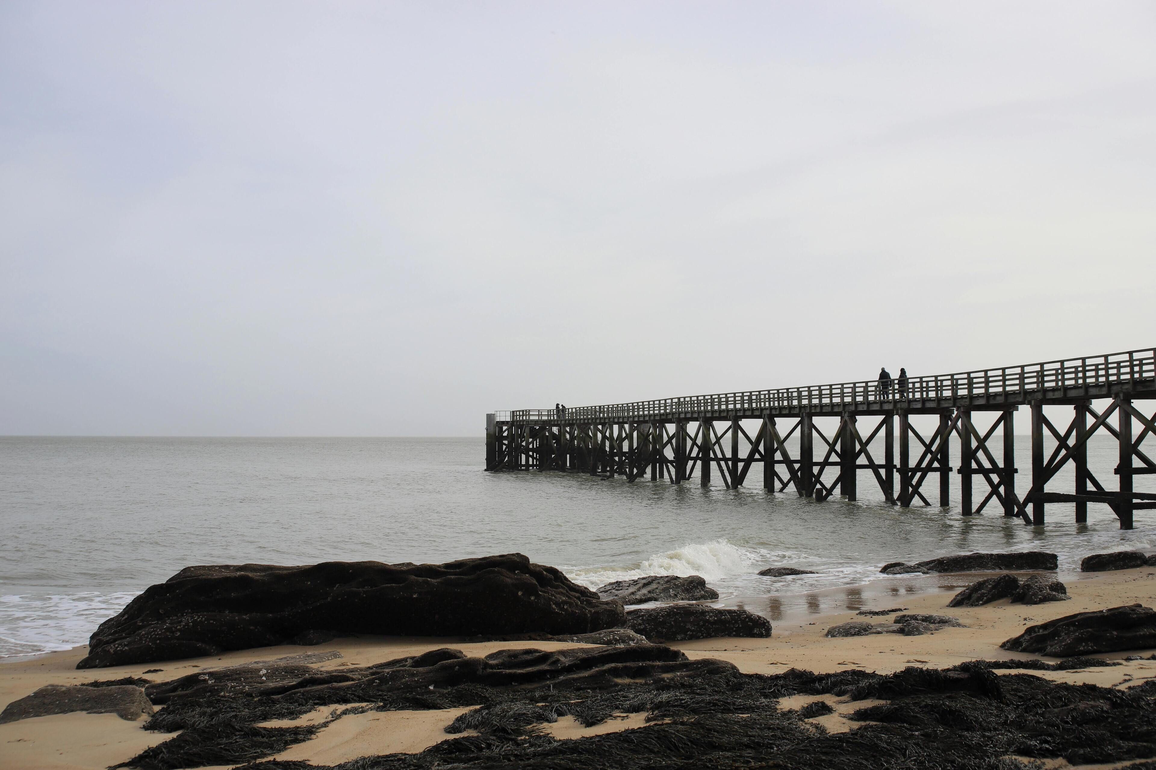 Un muelle se adentra en aguas tranquilas, rodeado de rocas y una playa de arena.