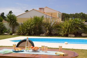 Une table au bord de la piscine, avec des fruits, des verres et un livre, entourée d’une végétation luxuriante et avec une maison en arrière-plan.
