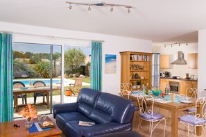 A living room with a blue leather sofa, dining table with chairs, a bookshelf, and a view of a swimming pool and garden.
