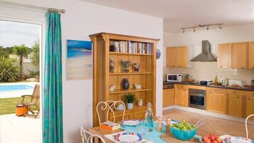 Una cocina moderna con armarios de madera, un microondas integrado y un horno. Hay una zona de comedor con una mesa preparada para dos, una estantería y vistas a una piscina exterior.