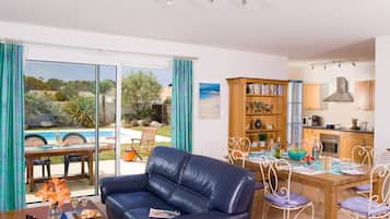 Ein Wohnzimmer mit blauem Ledersofa, einem Esstisch mit Stühlen, einem Bücherregal und Blick auf einen Pool und Garten.