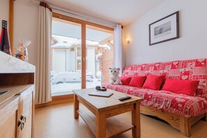 Ein gemütliches Wohnzimmer mit einem Holztisch, einem roten, gemusterten Sofa und Blick durch eine Glastür in einen verschneiten Außenbereich.