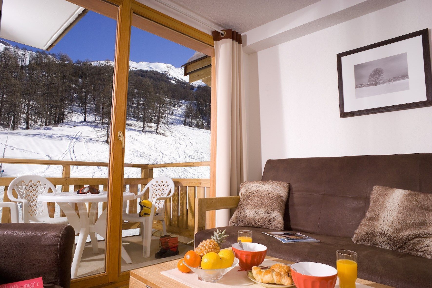 A cozy room with a view of a snowy mountain landscape. It includes a brown sofa, a dining table with breakfast items, and a balcony with a wooden railing.