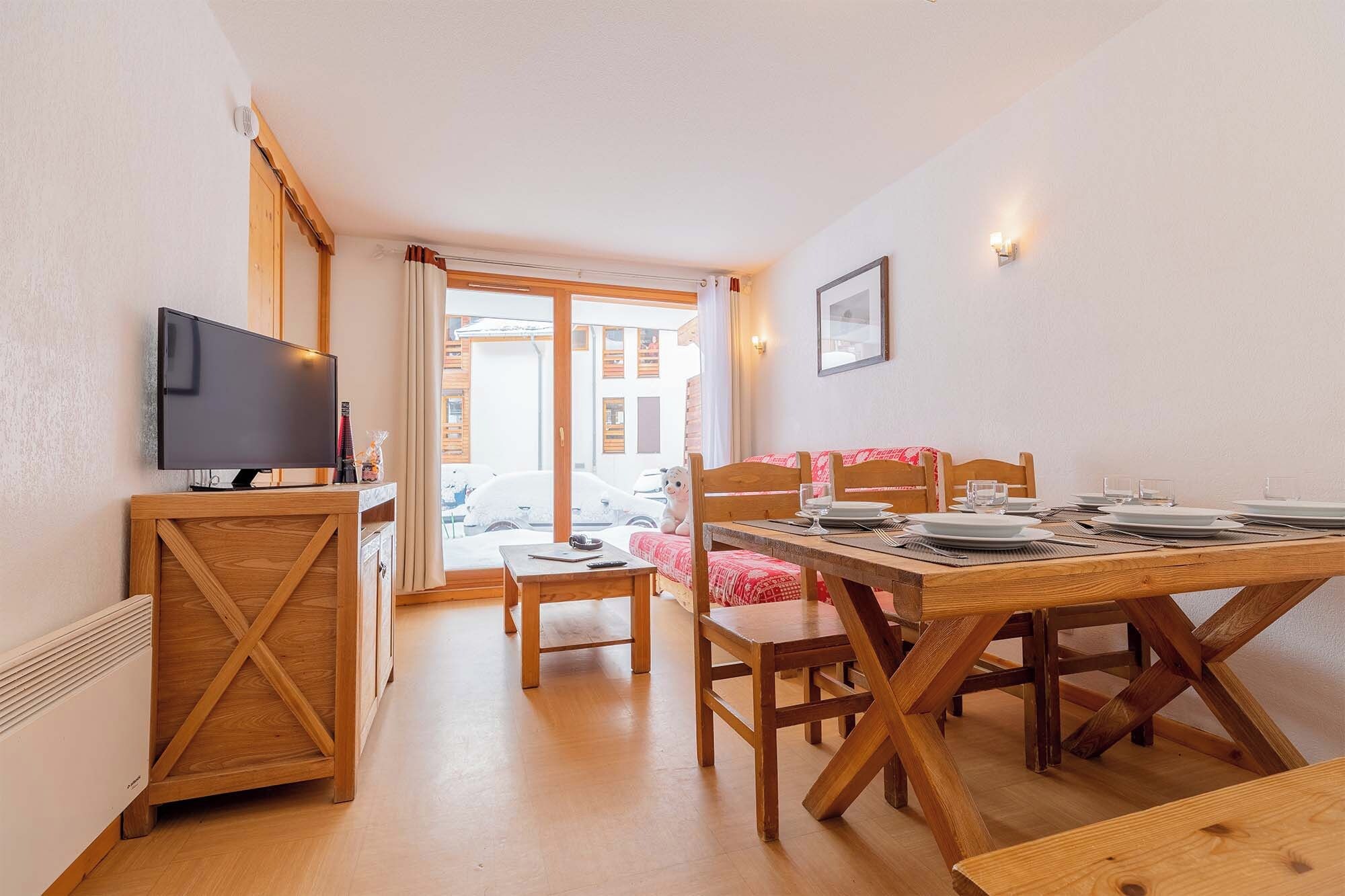 A dining area with a wooden table set for two, a television on a cabinet, and a view of the street outside.