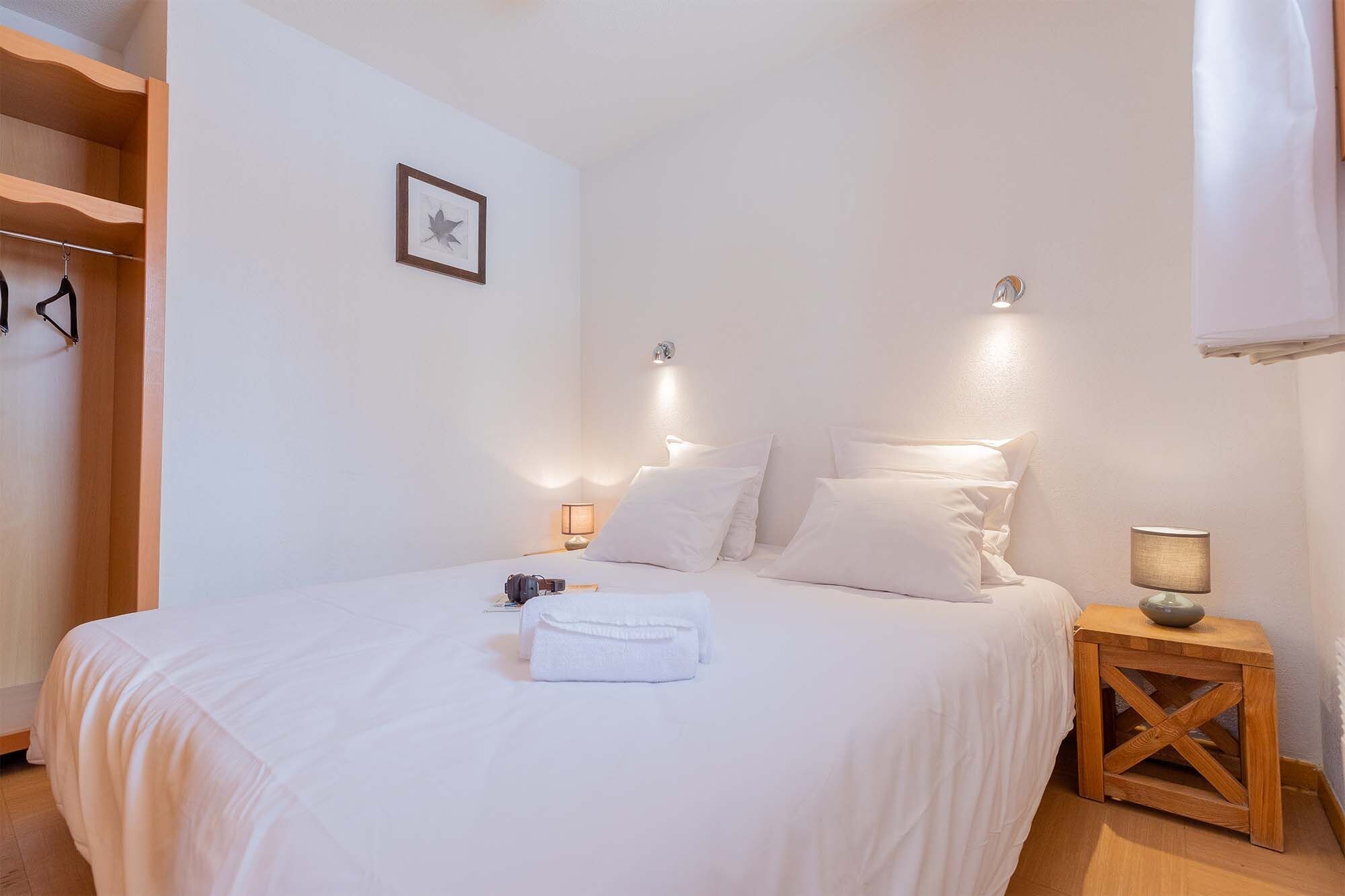 A neatly made bed with white linens, a towel, and a camera on the side. There are two bedside lamps and a framed picture on the wall.