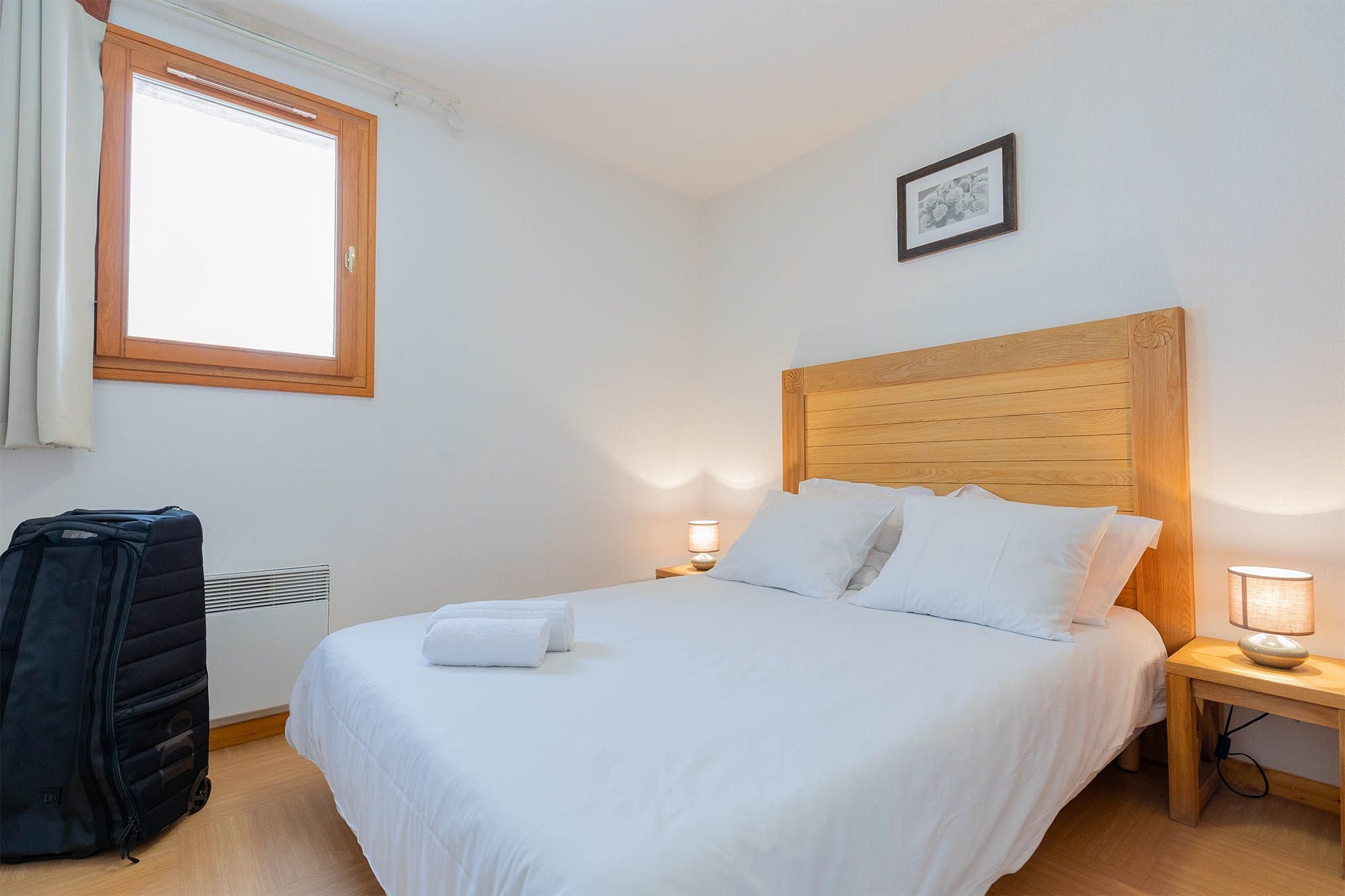 A hotel room with a wooden headboard, a bed with white linens, a window with curtains, a bedside lamp, and a suitcase.