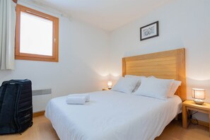 Ein Hotelzimmer mit Holz-Kopfteil, einem Bett mit weißen Leinen, einem Fenster mit Vorhängen, einer Nachttischlampe und einem Koffer.