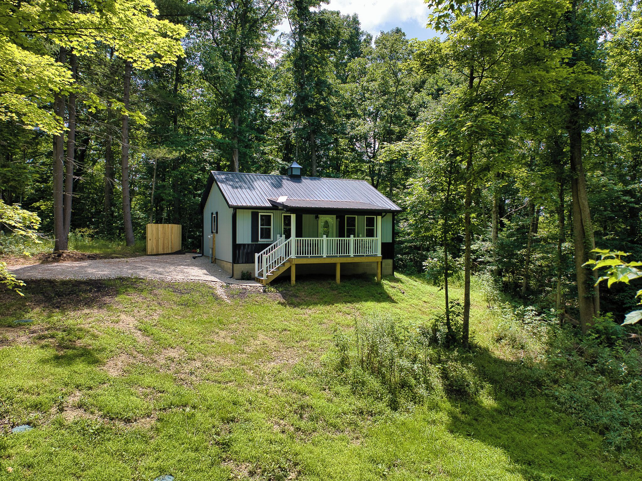 A small, single-story cabin with a porch, surrounded by dense forest.