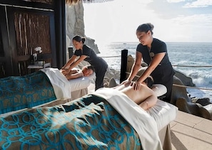 Ein Bereich für Massagetherapie mit zwei Massageliegen, eine wird von einem Klienten genutzt, die andere von einem Therapeuten. Der Raum bietet Blick auf den Ozean und eine felsige Küstenlinie.