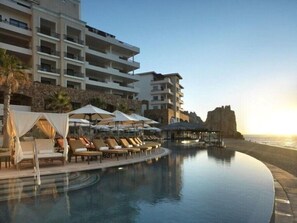 Ein Resort-Pool mit Liegestühlen und Sonnenschirmen, im Hintergrund ein Strand und eine felsige Klippe.