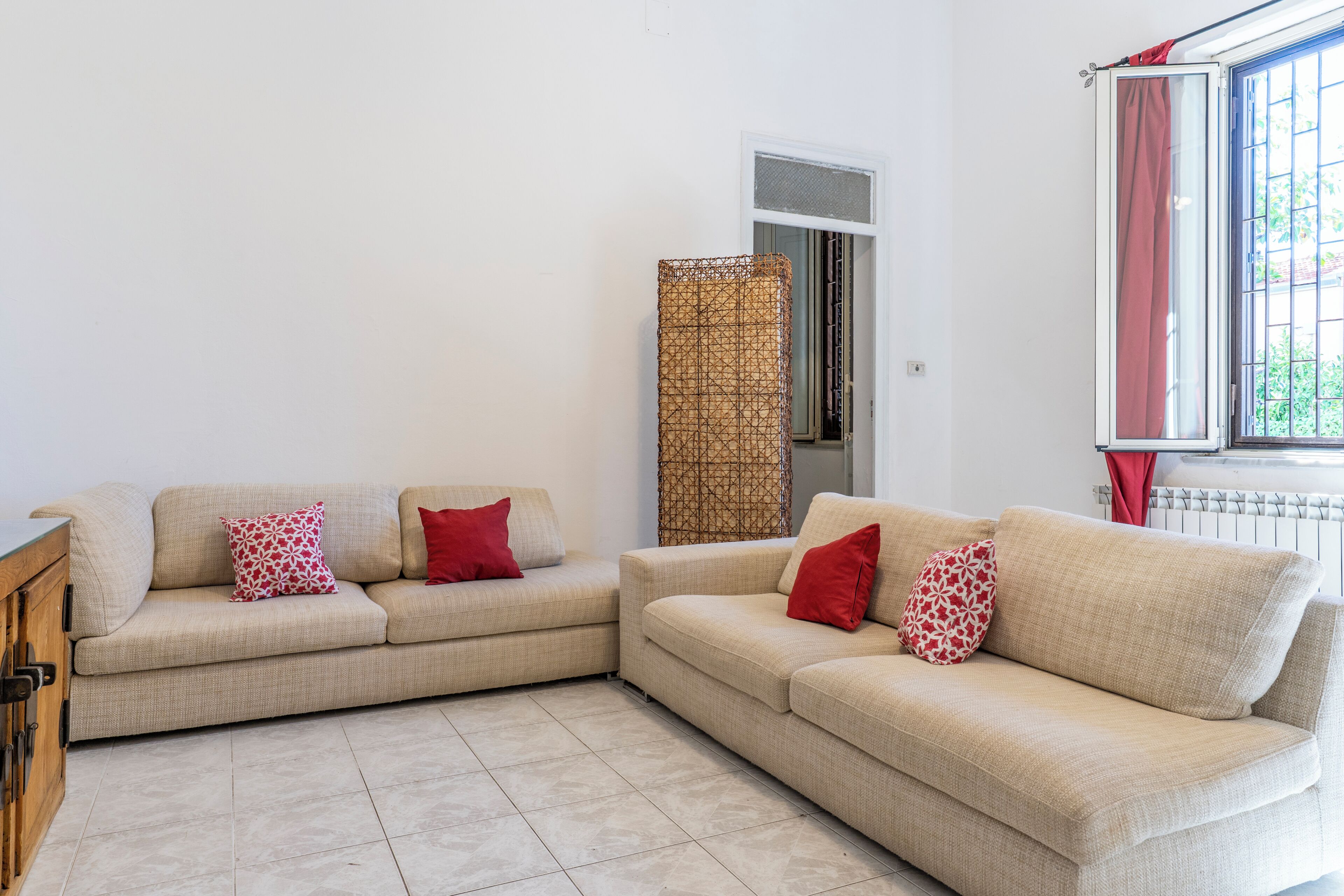 Ein Wohnzimmer mit zwei beigefarbenen Sofas, jedes mit Kissen in Rot und Weiß, einem Holzschrank, einem gemusterten Vorhang und einem Fenster mit Blick auf Grünflächen.