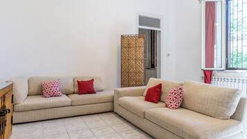Ein Wohnzimmer mit zwei beigefarbenen Sofas, jedes mit Kissen in Rot und Weiß, einem Holzschrank, einem gemusterten Vorhang und einem Fenster mit Blick auf Grünflächen.