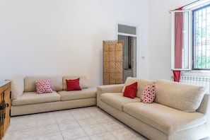 Ein Wohnzimmer mit zwei beigefarbenen Sofas, jedes mit Kissen in Rot und Weiß, einem Holzschrank, einem gemusterten Vorhang und einem Fenster mit Blick auf Grünflächen.