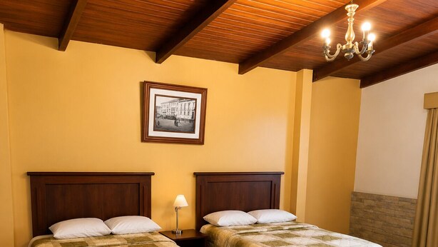 Habitación de hotel con dos camas, cabeceros de madera, una araña y un cuadro en la pared, además de un ventanal con cortinas.