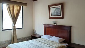 Un dormitorio con cama, mesita de noche, cuadro en la pared y ventana con cortinas.