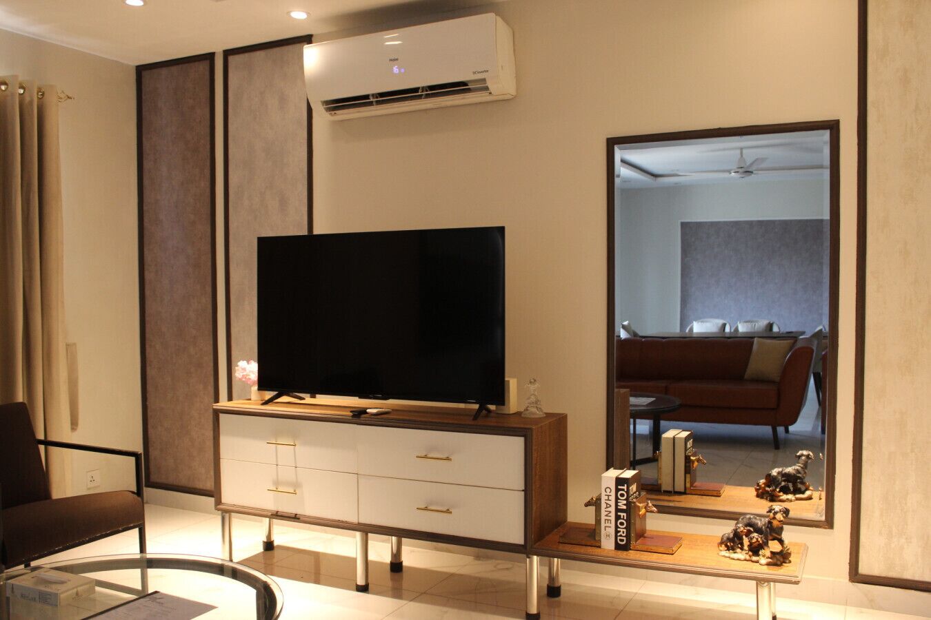 A modern living room with a flat-screen TV on a wooden console, a glass coffee table, and a large mirror reflecting the room.