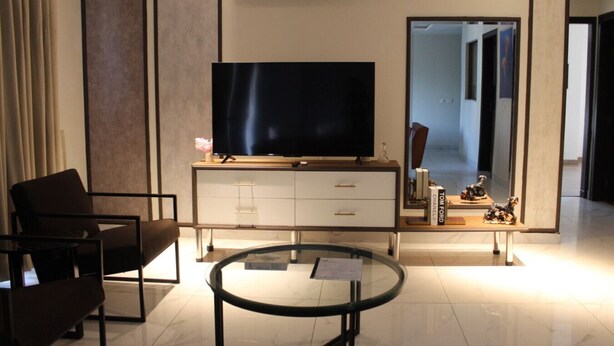 Ein modernes Wohnzimmer mit Flachbildfernseher, Glastisch, Sofa, einer Kommode mit Schubladen und einem großen Spiegel, der den Raum reflektiert.