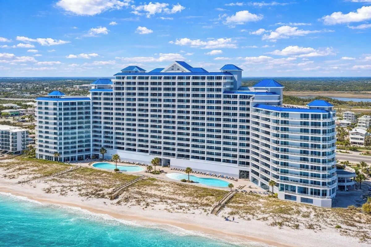 A large hotel with multiple floors and balconies, situated by a beach with clear blue waters.