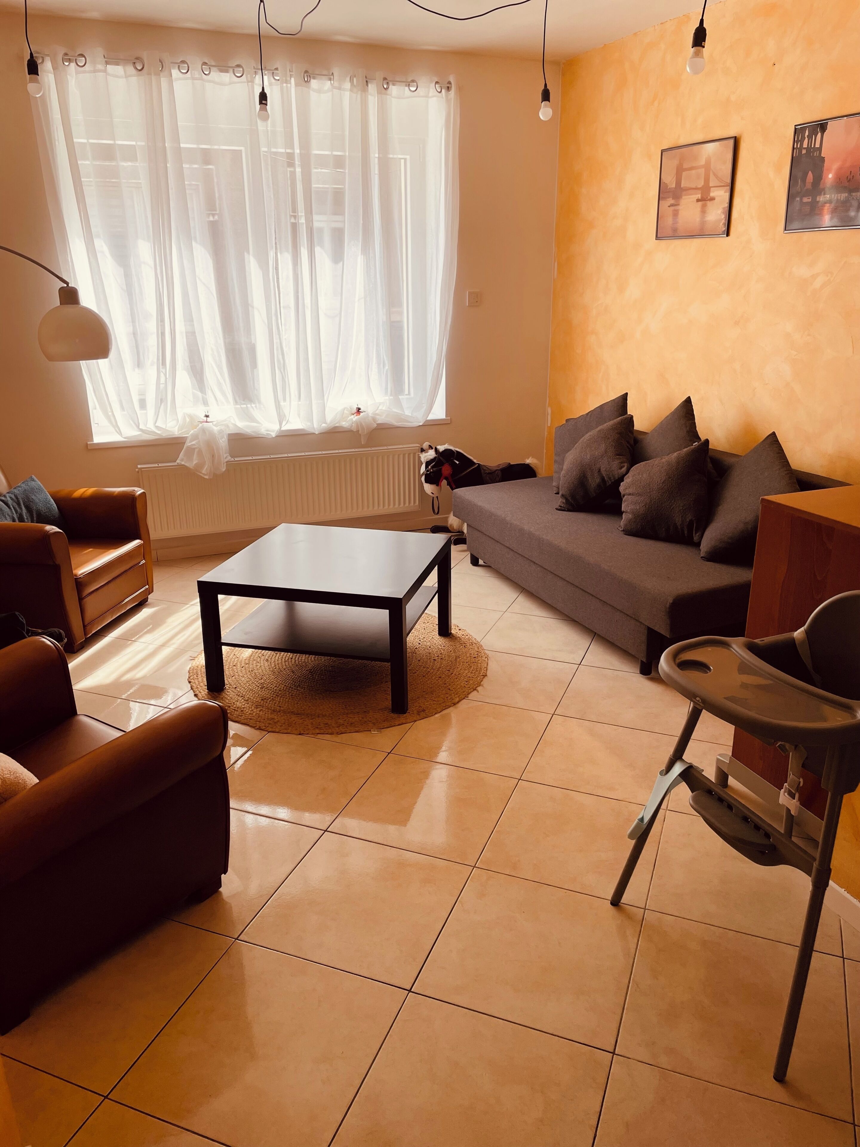 Un salón con un sofá, una mesa de centro y una silla. Hay una ventana con cortinas traslúcidas y un radiador debajo.