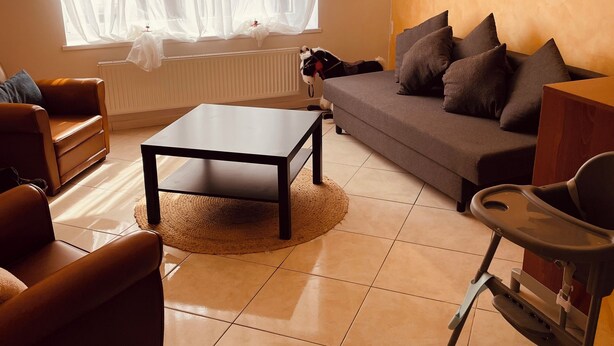 En stue med en sofa, et sofabord og en lænestol. Der er et vindue med lette gardiner og en radiator nedenunder.