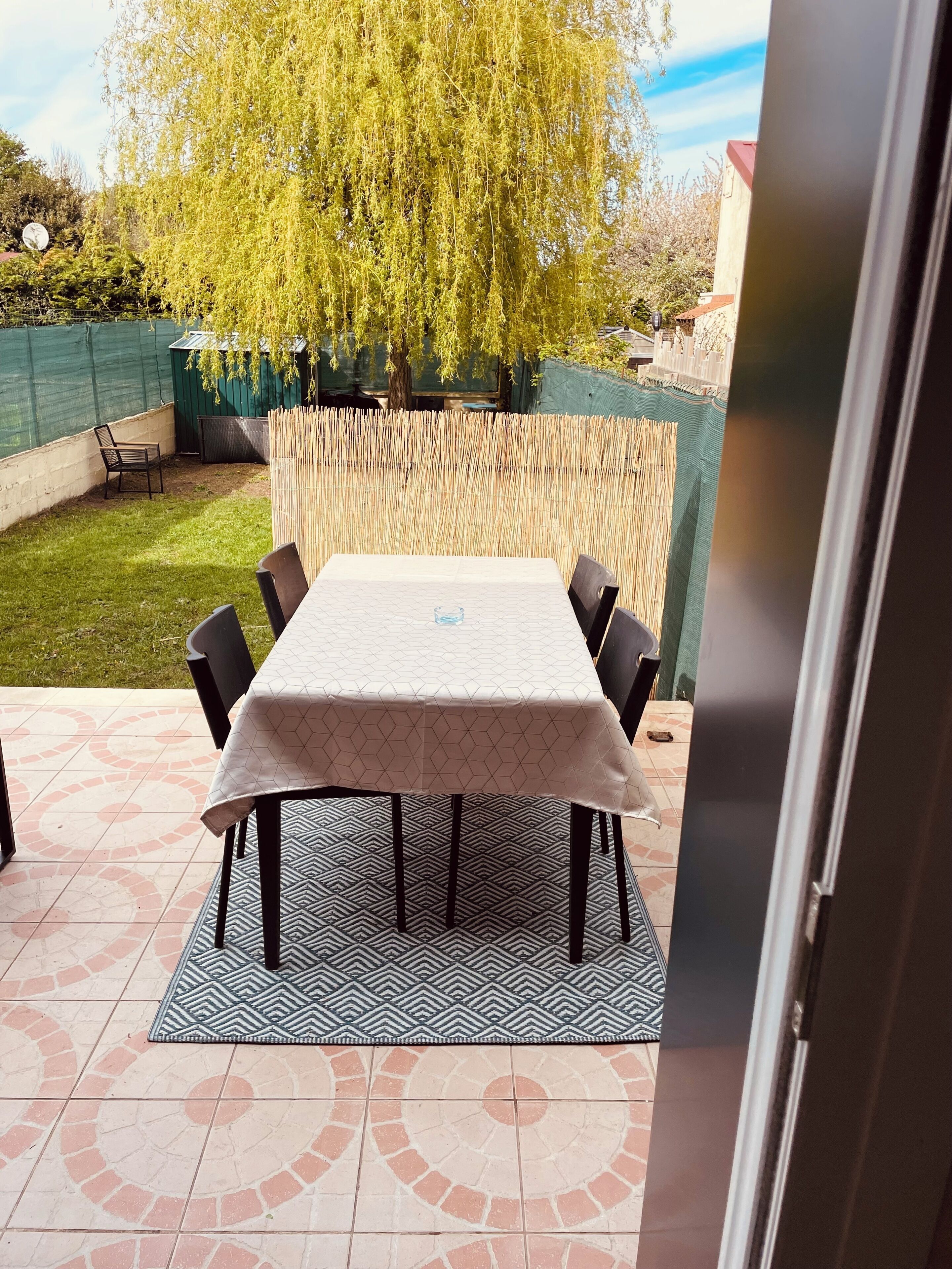 An outdoor patio with a table and chairs, surrounded by a bamboo fence, a green netted area, and a tree.