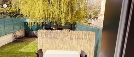 An outdoor patio with a table and chairs, surrounded by a bamboo fence, a green netted area, and a tree.