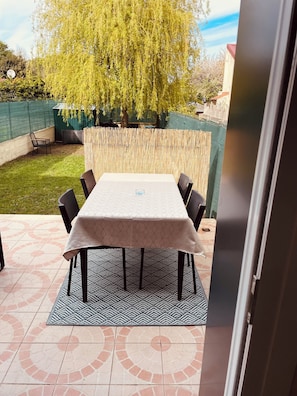 Een terras buiten met een tafel en stoelen, omgeven door een bamboomschutting, een groen gaasgedeelte en een boom.