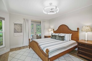 A bedroom with a wooden bed, a nightstand, a lamp, a window with curtains, and a view of greenery outside.