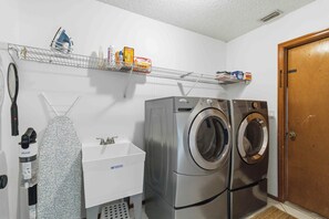 A laundry room with a washing machine, dryer, and a shelf with cleaning supplies. - Creekside Manor 1-Acre Luxury Home with Heated Pool near Lakewood Ranch (Bradenton)