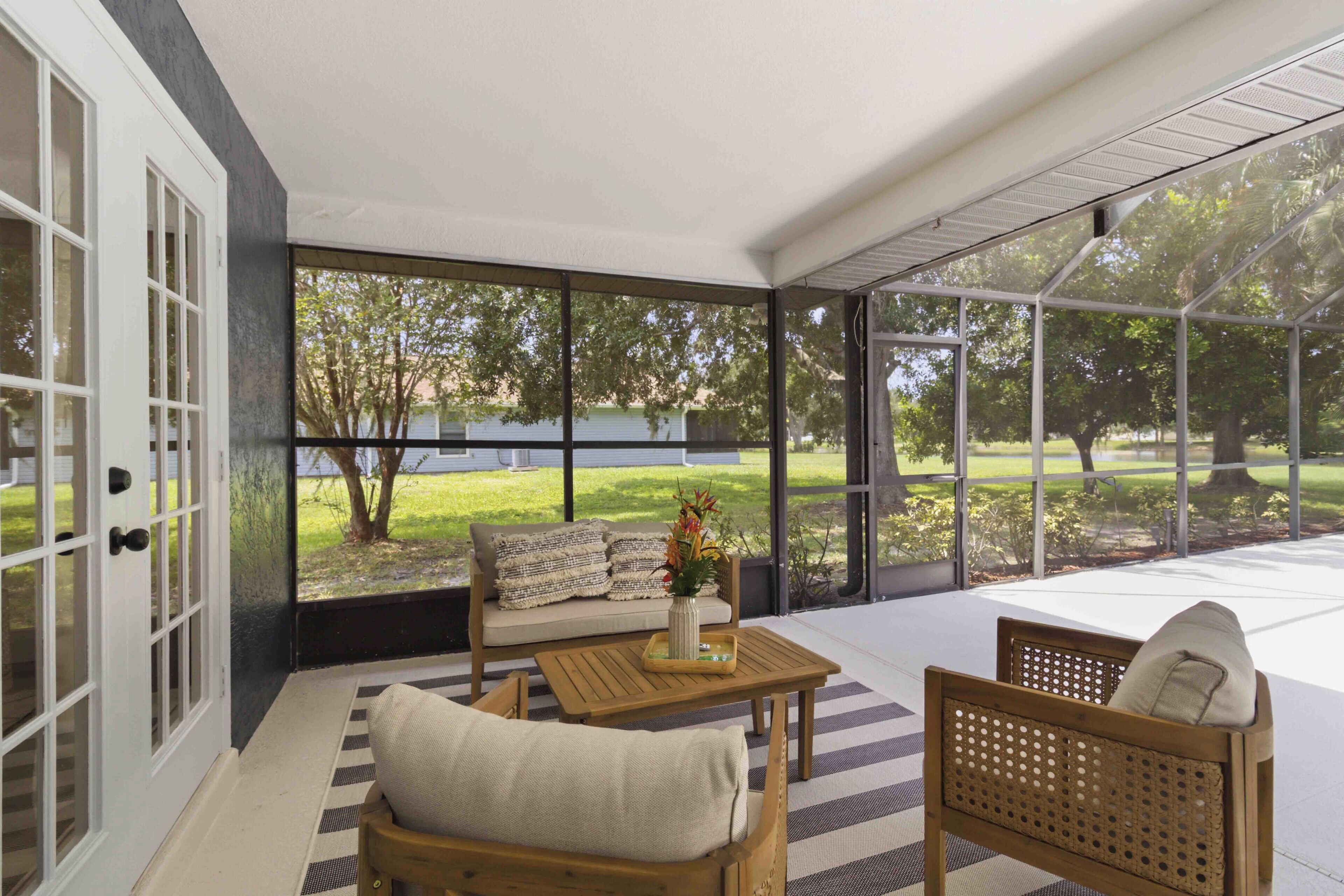 A sunlit room with a sofa, a wooden table, and a chair, featuring large windows and a view of a grassy area.