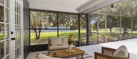 A sunlit room with a sofa, a wooden table, and a chair, featuring large windows and a view of a grassy area.