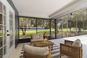 A sunlit room with a sofa, a wooden table, and a chair, featuring large windows and a view of a grassy area.