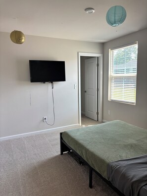 Une chambre à coucher avec un lit, un téléviseur fixé au mur, une porte donnant sur une autre pièce et une fenêtre avec des stores.