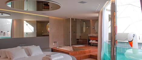 Ein modernes Hotelzimmer mit einem runden Bett, einem durch eine Glasscheibe abgetrennten Badezimmer und Blick aufs Meer.