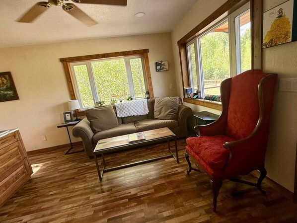 A room with a wooden floor, a sofa, a red chair, a table, and a window with a view of greenery.