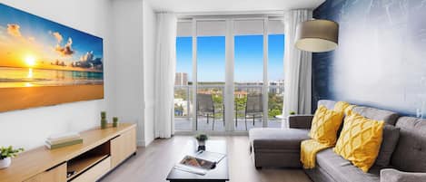 Un salon moderne avec une grande fenêtre, un canapé, une table basse et un tableau représentant un coucher de soleil.