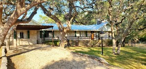 A stone house with a covered porch and a metal roof, surrounded by trees and a gravel driveway.