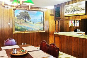 Ein Essbereich mit Holztisch, Stühlen, einem Landschaftsbild und einer Küchenzeile mit Spüle und Toaster.