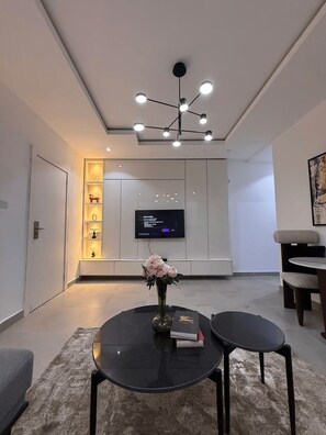 A modern living room with a round black table, a TV mounted on the wall, and a unique ceiling light fixture. - Perfect 1-Bedroom in Victoria Island with Pool, Fast Wi-Fi & 24/7 Power (Lagos)