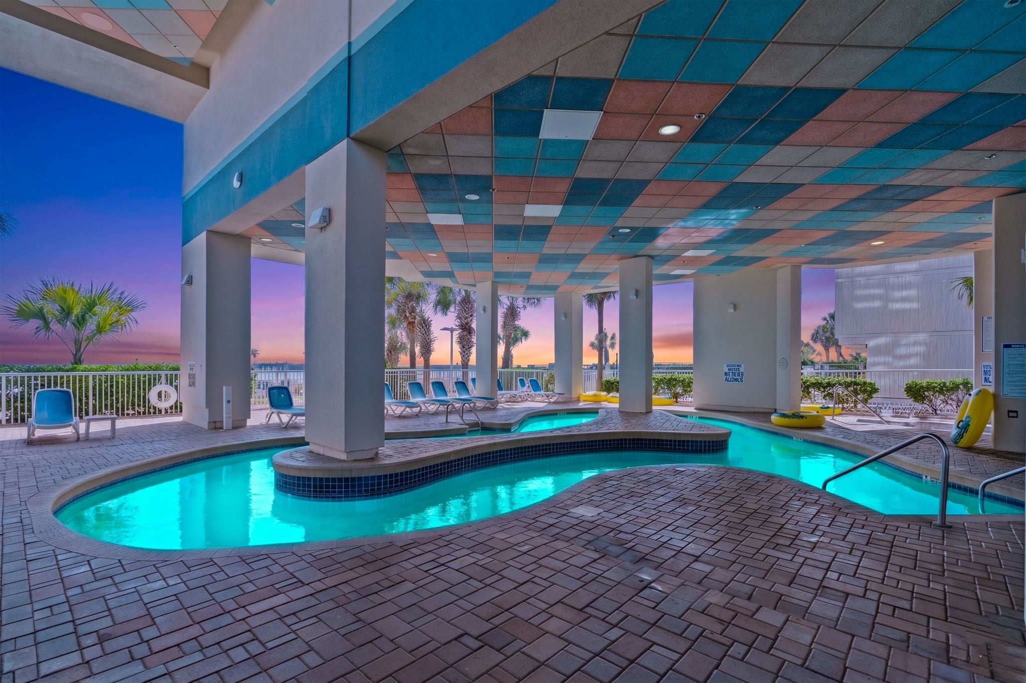An indoor swimming pool with tiled flooring, lounge chairs, and a view of palm trees at sunset.