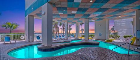An indoor swimming pool with tiled flooring, lounge chairs, and a view of palm trees at sunset.