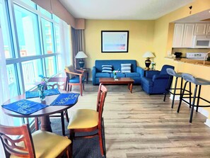 A living room with a sofa, coffee table, and chairs, a dining area with a table and chairs, and a kitchen with a counter and stools. - Carolinian Resort  Ocean View Condo (Myrtle Beach)