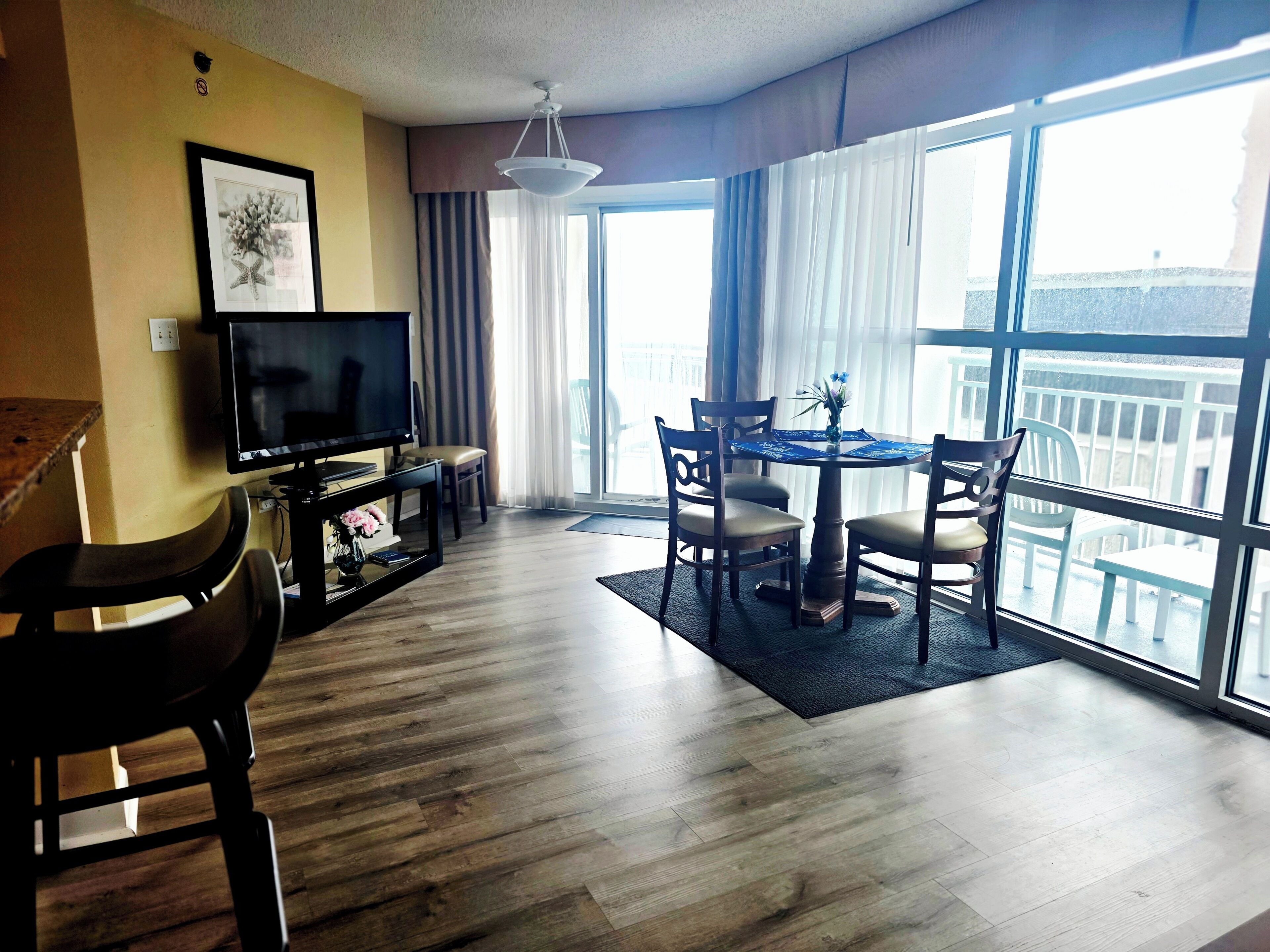 A hotel room with a television, a dining table with chairs, a balcony with a railing, and a large window.