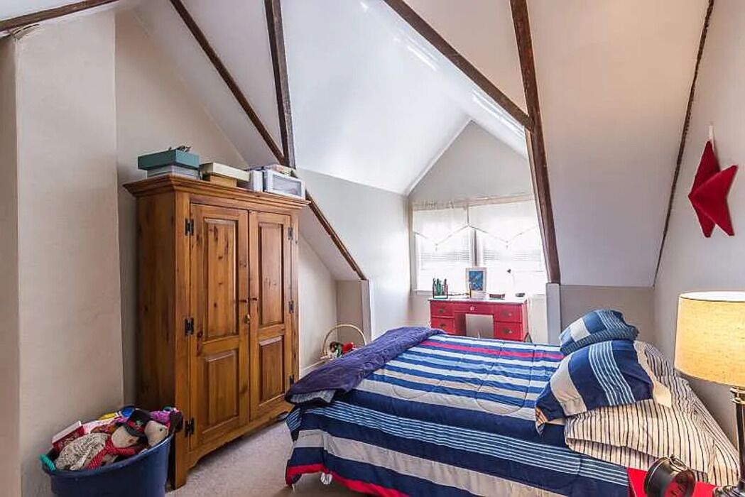A bedroom with a sloped ceiling, a wooden wardrobe, a bed with striped bedding, a red desk, and a lamp.