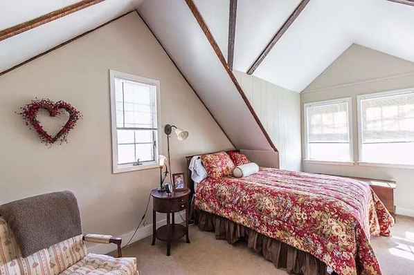 A bedroom with a sloped ceiling, a bed with a floral bedspread, a heart-shaped decoration on the wall, and a chair.