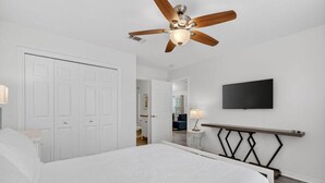Ein Schlafzimmer mit einem Bett, einem Deckenventilator, einem an der Wand befestigten Fernseher und einem Nachttisch mit Lampe.