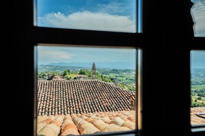 Une vue à travers une fenêtre sur un toit recouvert de tuiles, un clocher et un paysage avec des montagnes à l’horizon.