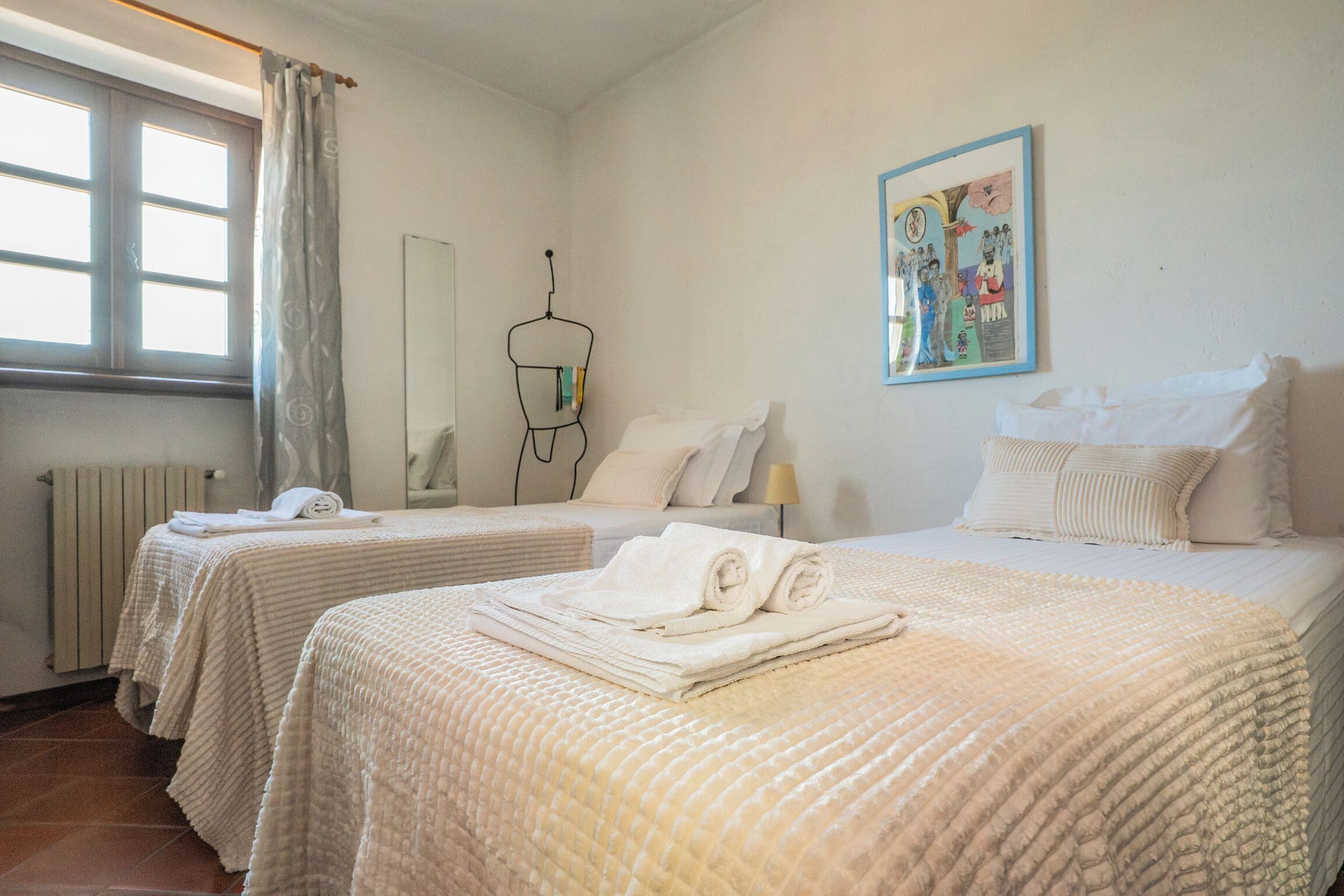 A hotel room with two beds, a radiator, a window with curtains, a wall-mounted towel rack, and a framed picture.