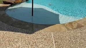 A swimming pool with a stone edge, a rock feature, and a blue umbrella. - Gig'em Getaway Pool House - Sleeps 10 (Bryan)
